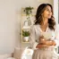A business-casual woman stands in a bright, modern, plant-filled living space, holding a journal and pen. She is looking out a window thoughtfully, potentially reflecting on career choices or personal goals.