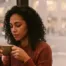 A close-up photograph of a contemplative woman with curly hair holding a warm beverage, looking out a window at a blurred, rainy Manhattan cityscape.